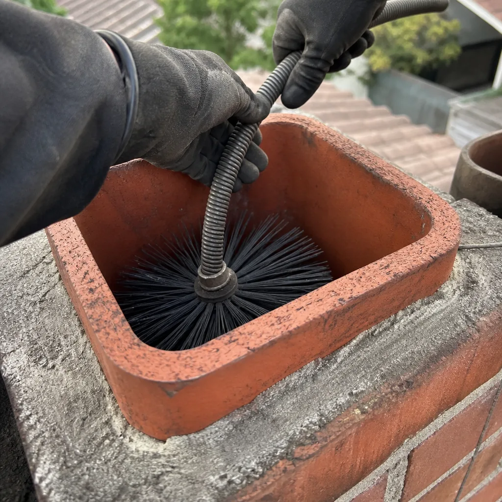Professional chimney sweep cleaning a brick chimney flue with a wire brush.